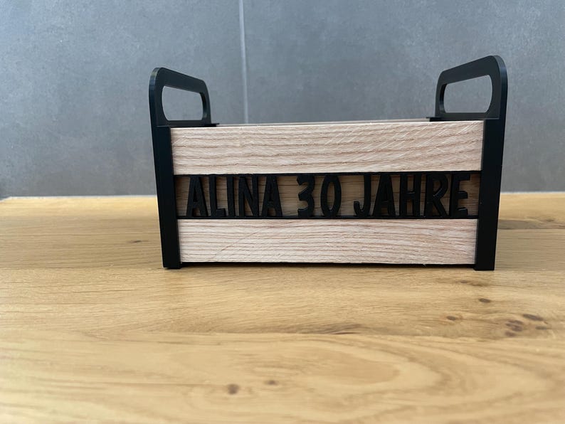 May include: A wooden box with black handles and trim. The front of the box features the text "ALINA 30 JAHRE" in black, cut-out letters. The box has a light wood grain finish and is sitting on a wooden surface.