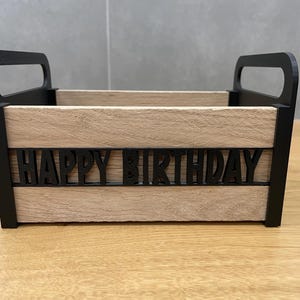 May include: A wooden box with black handles and the words "HAPPY BIRTHDAY" cut out on the front. The box has a light brown, wood-grain appearance and is designed for gift-giving or decorative purposes.