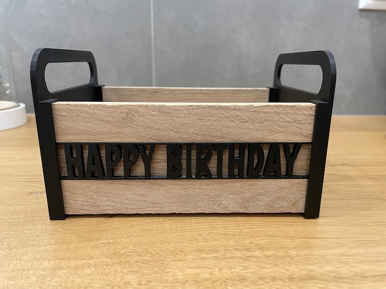 May include: A wooden box with black handles and the words "HAPPY BIRTHDAY" cut out on the front. The box has a light brown, wood-grain appearance and is designed for gift-giving or decorative purposes.