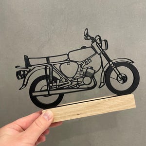 Op de afbeelding: Een zwart, plat, tweedimensionaal silhouet van een motorfiets gemonteerd op een rechthoekige, lichtgekleurde houten voet. Het ontwerp van de motorfiets toont gedetailleerde contouren van het frame, de wielen, de motor, het zadel en het stuur.