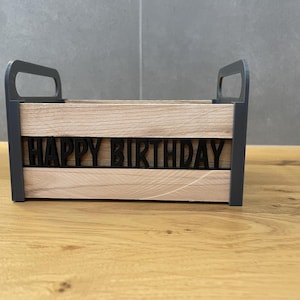 May include: A wooden box with a black frame and handles. The front of the box features the words "HAPPY BIRTHDAY" in black, cut-out letters. The box is made of light-colored wood and sits on a wooden surface.