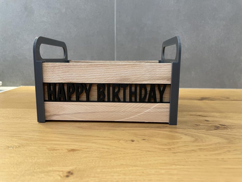 May include: A wooden box with a black frame and handles. The front of the box features the words "HAPPY BIRTHDAY" in black, cut-out letters. The box is made of light-colored wood and sits on a wooden surface.