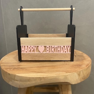 Könnte beinhalten: Ein rechteckiger Holzkorb mit hellen Holzpaneelen, schwarzen Seitenstützen und einem Griff aus Naturholz. Auf der Vorderseite befindet sich der hellrosa Schriftzug "HAPPY BIRTHDAY" mit einem Herzsymbol zwischen den beiden Wörtern.