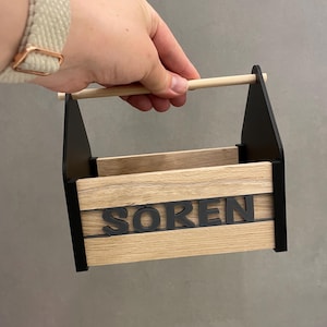 Könnte beinhalten: Ein kleiner rechteckiger Aufbewahrungskorb mit hellen Holzmaserungs-Seitenwänden und mattschwarzen Endstützen. Ein zylindrischer Griff aus hellem Holz überspannt die Oberseite. Der Name SOREN ist in erhabenen schwarzen Großbuchstaben vorne angebracht.