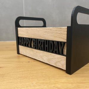 May include: A wooden box with black handles and sides. The front of the box has the words "HAPPY BIRTHDAY" cut out in black. The box is sitting on a wooden surface.