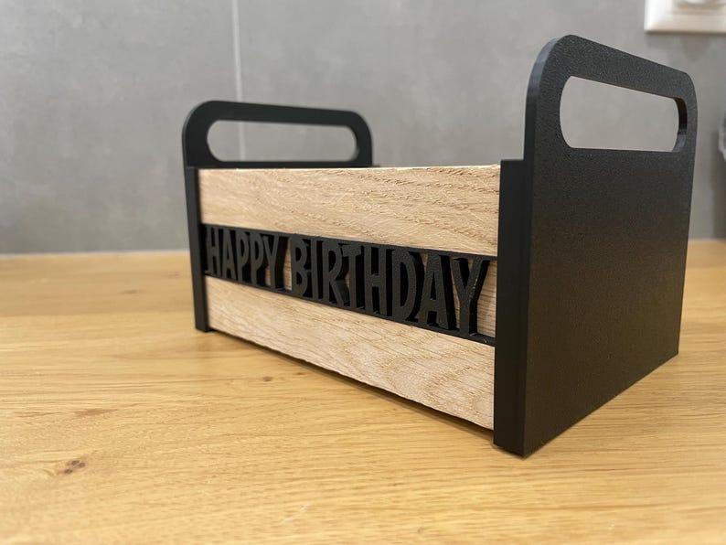 May include: A wooden box with black handles and sides. The front of the box has the words "HAPPY BIRTHDAY" cut out in black. The box is sitting on a wooden surface.