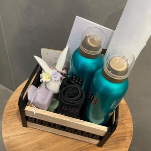 May include: A gift basket with teal-colored bottles, a black rolled item, a purple candle, and a decorative bunny. The basket has the words "BESTE MAMA" on the front. The items are arranged on a wooden table.
