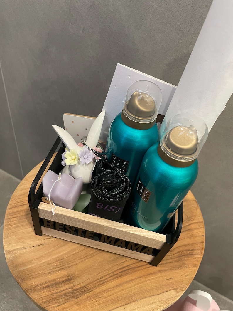 May include: A gift basket with teal-colored bottles, a black rolled item, a purple candle, and a decorative bunny. The basket has the words "BESTE MAMA" on the front. The items are arranged on a wooden table.