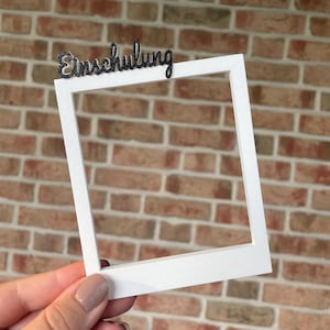 Könnte beinhalten: Ein weißer, quadratischer Fotorahmen im Stil eines Sofortbildfotos mit dem Wort "Einschulung" in schwarzer, glitzernder Schreibschrift am oberen Rand. Der Rahmen hat eine matte Oberfläche und einen breiten unteren Rand, der an ein Foto erinnert.