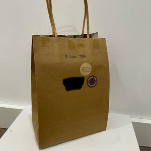 Puede incluir: Una bolsa de papel marrón con asas de papel retorcido. La bolsa tiene el texto "B-Wave Tute" y varias pegatinas circulares. Una pegatina dice "handmade with love" y otra dice "thank you".