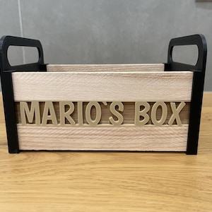 May include: A wooden box with black handles and trim, featuring the text "MARIO'S BOX" in gold lettering. The box has a natural wood grain appearance and is designed for storage or display.