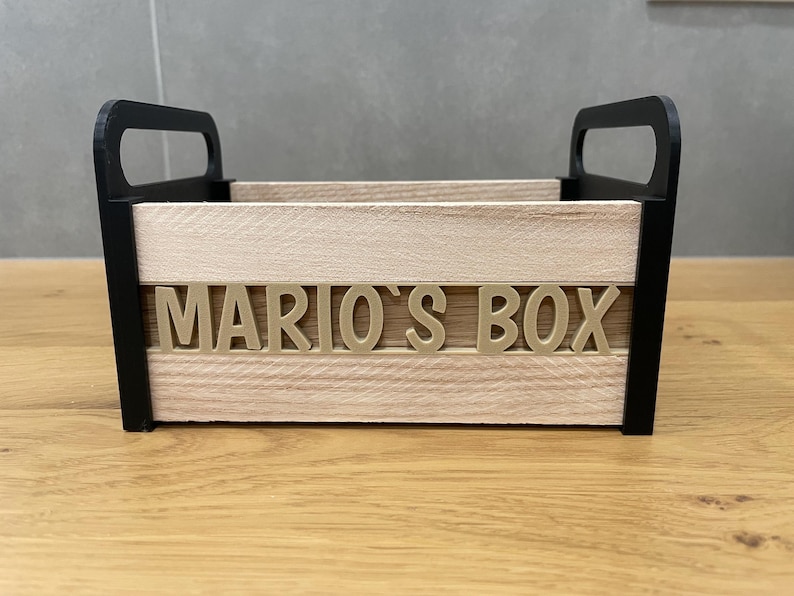 May include: A wooden box with black handles and trim, featuring the text "MARIO'S BOX" in gold lettering. The box has a natural wood grain appearance and is designed for storage or display.