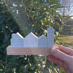 Könnte beinhalten: Eine Miniatur-Stadtlandschaftsskulptur mit vier hellblauen Gebäuden auf einem Holzsockel. Die Gebäude umfassen ein Haus, eine Kirche, ein weiteres Haus und ein hohes Gebäude. Die Skulptur wird in einer Hand vor einem Hintergrund aus grünem Laub gehalten.