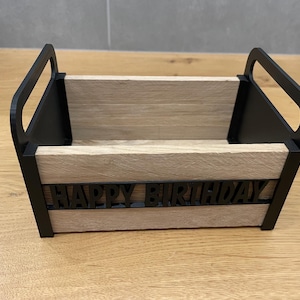 May include: A wooden box with black handles and trim, featuring the words "HAPPY BIRTHDAY" in black letters. The box has a light brown, wood-grain appearance and is designed for decorative or gift-giving purposes.