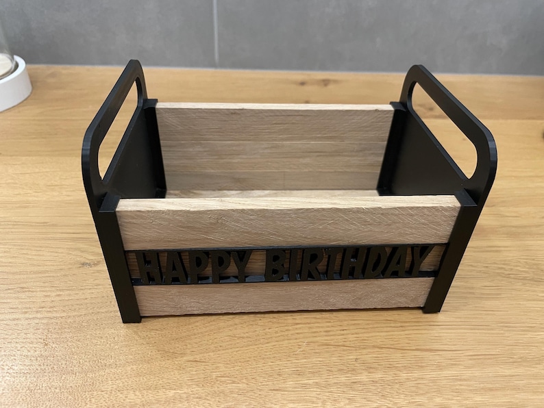 May include: A wooden box with black handles and trim, featuring the words "HAPPY BIRTHDAY" in black letters. The box has a light brown, wood-grain appearance and is designed for decorative or gift-giving purposes.