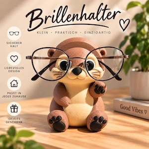 May include: A brown and cream-colored otter-shaped eyeglass holder featuring a pair of black-rimmed glasses resting on its face. The text Brillenhalter, Klein, Praktisch, Einzigartig, Sicherer Halt, Liebevolles Design, Passt in jedes Zuhause, Ideales Geschenk, and Good Vibes are visible.