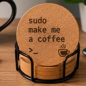Könnte beinhalten: Ein Stapel runder, hellbrauner Korkuntersetzer in einem schwarzen Metalldrahtständer. Der oberste Untersetzer zeigt den schwarzen Aufdruck "sudo make me a coffee", gefolgt von einem Befehlszeilensymbol und einem kleinen Symbol einer dampfenden Kaffeetasse.