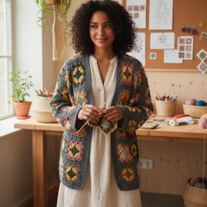 Könnte beinhalten: Ein handgefertigter, offener Häkelcardigan im Granny-Square-Muster. Der Cardigan hat eine graue Basis mit bunten Blumenquadraten in Gelb-, Rosa- und Grüntönen. Die Frau hält Häkelnadeln und ein teilweise fertiggestelltes Quadrat.