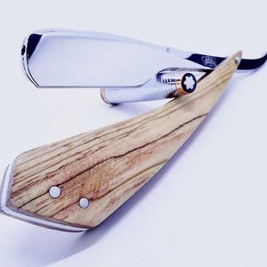 Puede incluir: Una navaja de afeitar con una hoja de metal plateado pulido y un mango de madera marrón claro con vetas visibles. El mango está asegurado con dos remaches plateados y el punto de pivote muestra un pequeño emblema de estrella en blanco y negro.