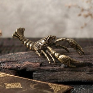 Puede incluir: Una pequeña figura de langosta de metal color latón con un cuerpo segmentado y texturizado y dos pinzas delanteras prominentes. La escultura presenta detalles intrincados en el caparazón y las extremidades, capturando la forma del crustáceo.