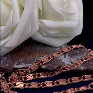 May include: A copper-coloured chain necklace with a distinctive S-link design, displayed on a textured stone surface. A large white rose is in the background, with a dark blue backdrop.