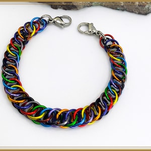 Pode incluir: Uma pulseira colorida de malha de corrente com um arco-íris de tons, incluindo vermelho, laranja, amarelo, verde, azul e roxo. A pulseira é feita de pequenos anéis de metal interligados e tem um fecho prateado.