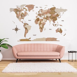 May include: A wooden world map with country outlines in brown and beige tones is mounted on a white wall. A pink sofa and a white rug are in front of the map. A compass is to the left of the map.