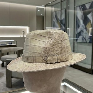 Puede incluir: Un sombrero fedora color crema con un patrón texturizado similar al de un cocodrilo. El sombrero presenta una banda a juego con una hebilla plateada. El ala es ancha y el sombrero se exhibe en una cabeza de maniquí.