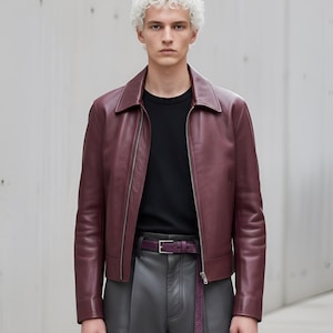 May include: A burgundy leather jacket with a zip closure, worn over a black crew neck shirt. The jacket is paired with gray leather pants and a matching burgundy belt. The outfit is a modern take on classic leather wear.