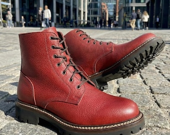 May include: A pair of red leather boots with a textured finish. The boots have a lace-up design with dark brown laces, a thick black sole, and a stitched welt. The boots are displayed on a cobblestone surface.