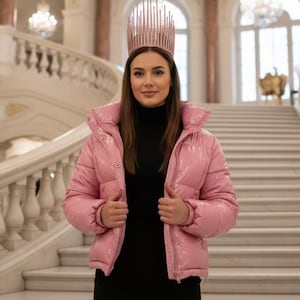 May include: A woman in a pink puffer jacket, black turtleneck, and a black dress, wearing a rose gold crown, stands on a grand staircase. The background features chandeliers and architectural details.