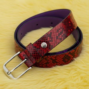 Può includere: Una cintura rossa con motivo a pelle di serpente con fibbia argentata e interno viola. La cintura è arrotolata su una superficie gialla e sfocata. La cintura ha un singolo borchia argentata vicino alla fibbia.