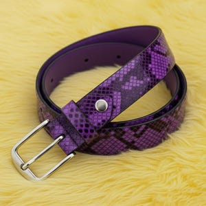 May include: A purple belt with a snakeskin pattern and a silver buckle. The belt is made of a purple material with a darker purple and black snakeskin design. The buckle is silver and rectangular.