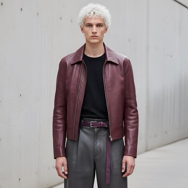 May include: A burgundy leather jacket with a zip closure, paired with a black crew neck shirt and gray leather pants. A matching burgundy belt with a silver buckle completes the outfit. The jacket has a classic collar.