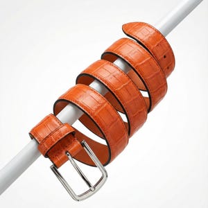 Op de afbeelding: Een oranje leren riem met krokodillenstructuur is om een witte paal gewikkeld. De riem heeft een zilverkleurige gesp en een zwarte binnenkant. De riem is een stijlvol accessoire.