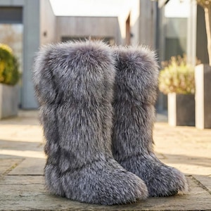Puede incluir: Un par de botas de piel grises. Las botas son hasta la rodilla y están hechas de piel larga y suave. Las botas están sobre una superficie de piedra. Las botas son de color neutro y se pueden usar en invierno.