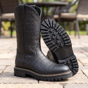 Könnte beinhalten: Ein schwarzer Lederstiefel mit texturiertem Krokodilleder-Muster, hohem Schaft, Zuglaschen und einer dicken, profilierten Gummisohle mit sichtbaren Nähten entlang des Rahmens, der sowohl das Seitenprofil als auch das Sohlenprofil zeigt.