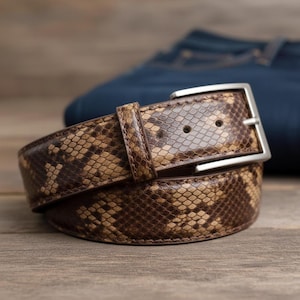 May include: A brown and tan snakeskin-patterned belt with a silver-colored buckle. The belt is made of leather and is rolled up. A pair of blue jeans is in the background.