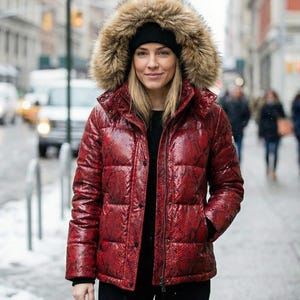 May include: A red puffer jacket with a snakeskin pattern and a fur-trimmed hood. The jacket is buttoned up and worn with a black beanie. The background shows a city street with people and buildings.