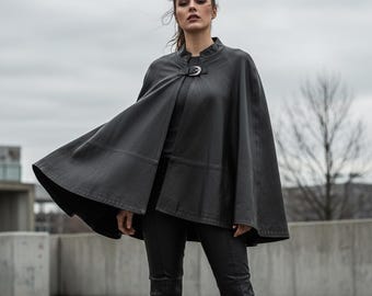 May include: A black leather cape with a buckle fastening at the neck. The cape is knee-length and worn with black trousers and boots. The model is standing outside on a cloudy day.