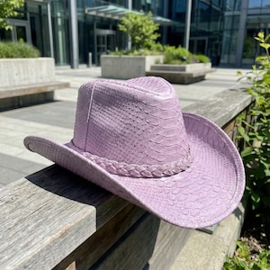 Può includere: Un cappello da cowboy color lavanda con una trama a pelle di serpente. Il cappello presenta una fascia intrecciata attorno alla corona e un'ampia tesa. Il cappello è appoggiato su una superficie di legno, con un edificio moderno sullo sfondo.