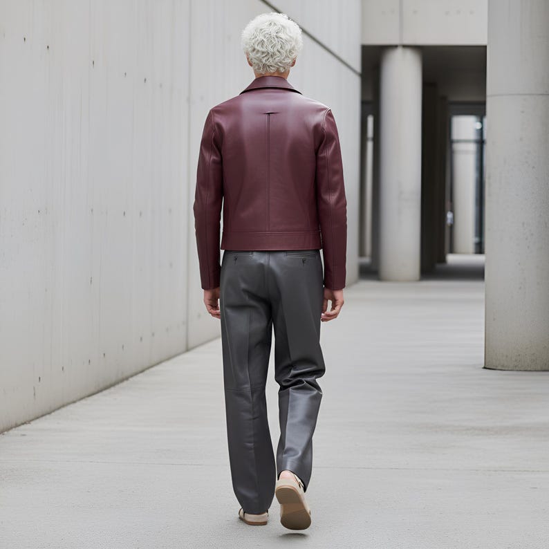 May include: A burgundy leather jacket with a classic collar and a tailored fit. The jacket is paired with gray trousers and beige sandals. The person is walking away in a modern architectural setting.