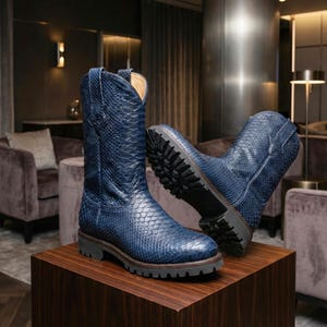 May include: A pair of blue cowboy boots with a textured, snakeskin-like pattern. The boots have a tall shaft, a rounded toe, and a lug sole. They are displayed on a dark wooden cube.