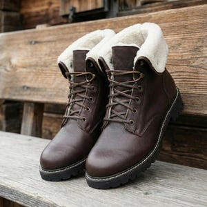 Close up front view of brown leather boots featuring soft white shearling lining and a padded ankle collar. These mens winter shoes display premium leather texture with visible grain and rounded toes. The black rubber outsoles have deep treads for winter grip and distinctive white perimeter stitching. Brown leather laces are tied through antique finish metal grommets. The boots sit on a weathered grey wood bench with a blurry alpine background. Authentic craftsmanship is evident in every detail.