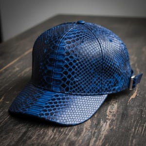 May include: A blue baseball cap with a snakeskin pattern. The cap features a curved brim and an adjustable strap with a silver buckle. The cap is made of a textured material and has a button on top.
