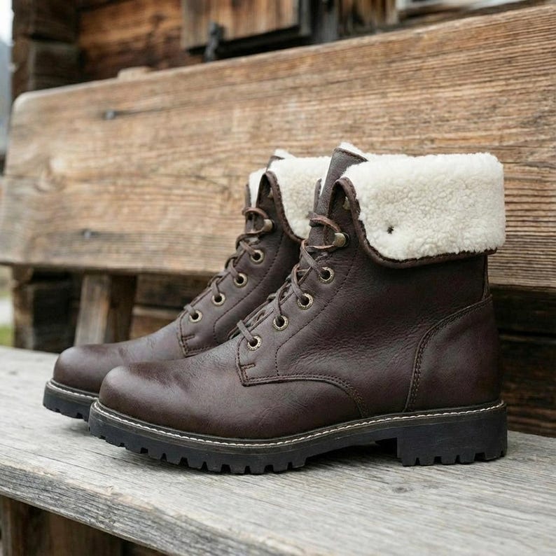 Side profile view of dark brown leather winter boots on a rustic wooden bench. This angle highlights the thick black lug sole and the white contrast stitching along the base. The plush white faux fur collar is folded down showcasing the inner warmth and insulation. The boot has a high top design with metallic eyelets for the brown laces. The background features a weathered wood wall and a glimpse of a mountain valley. These boots combine rugged outdoor functionality with a stylish urban look