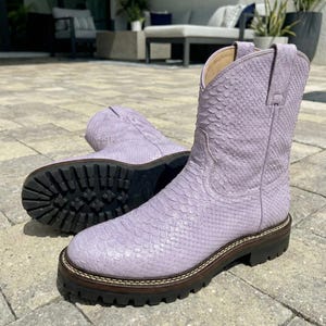 Puede incluir: Un par de botas de vaquero color lavanda con un estampado de piel de serpiente. Las botas tienen punta redonda, caña corta y suela gruesa. Las suelas son negras con ribete marrón. Las botas están sobre una superficie de piedra.