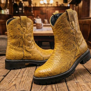 May include: A pair of yellow cowboy boots with a snakeskin pattern. The boots have a black sole and a short heel. The boots are made of a textured material and are sitting on a wooden surface.