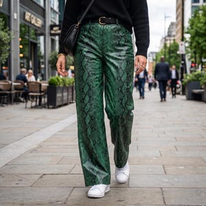 Puede incluir: Pantalones de talle alto con un vibrante estampado de piel de serpiente en verde y negro, combinados con un cinturón negro, un suéter negro de manga larga, un bolso de hombro negro y zapatillas blancas. El pantalón tiene un corte recto.