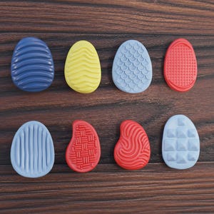 Könnte beinhalten: Acht farbenfrohe, eiförmige Objekte mit strukturierten Oberflächen sind auf einem dunklen Holzmaserungshintergrund angeordnet. Die Eier sind blau, gelb, hellblau und rot, mit verschiedenen Mustern. Ostermotiv.